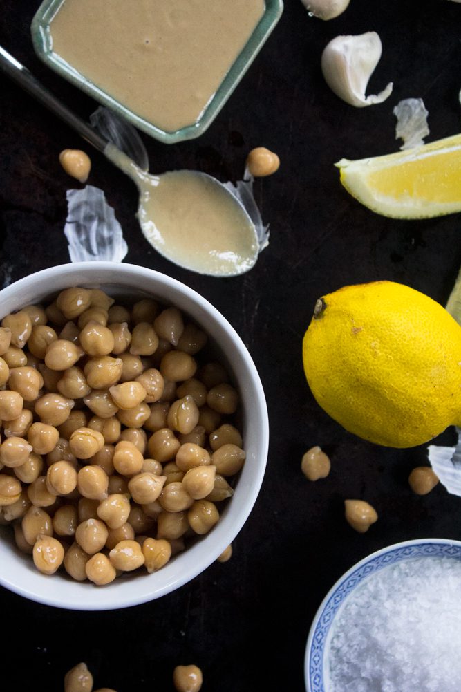 Ingredients for traditional hummus: garbanzo beans in a bowl, one lemon, tahini paste, spoon, sea salt, and one garlic clove