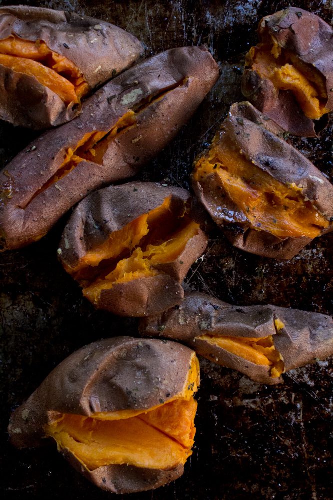Red skinned sweet potatoes on a baking sheet sprinkled with fresh thyme, rosemary, sea salt and fresh pepper cut open to cool after roasted