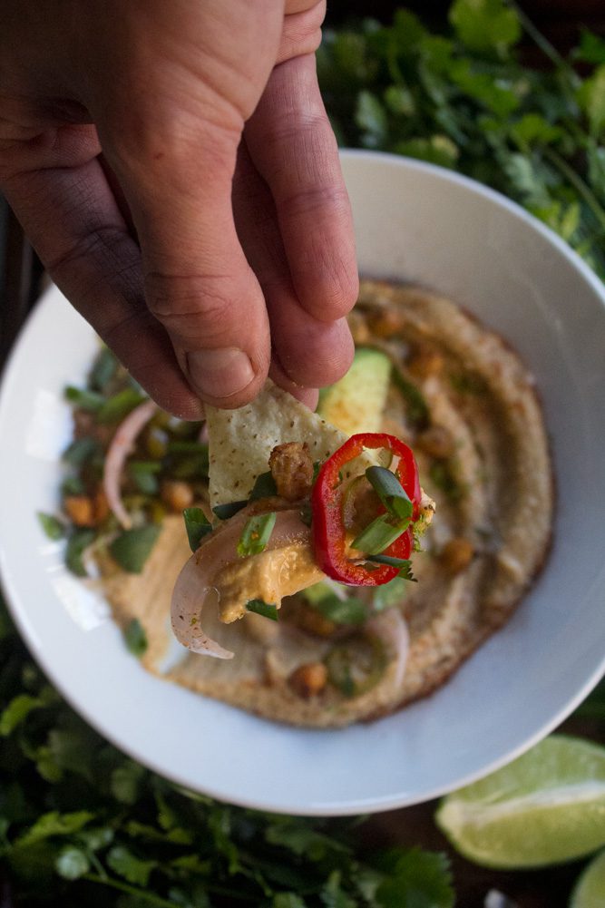Close-up of hand holding tortilla chip and Taco Hummus with pickled salsa, crispy chickpeas, cilantro, and avocado