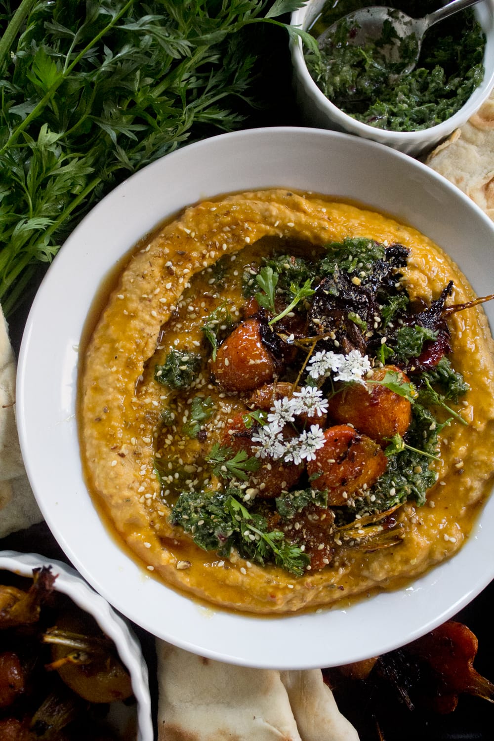 Plated dukkah and carrot hummus with zhoug - sweet, garden fresh carrots roasted with harissa, honey, and cumin seeds added to hummus and dressed with addiitonal roasted carrots, dukkah, zhoug, cilantro leaves + flowers, and drizzled with honey.